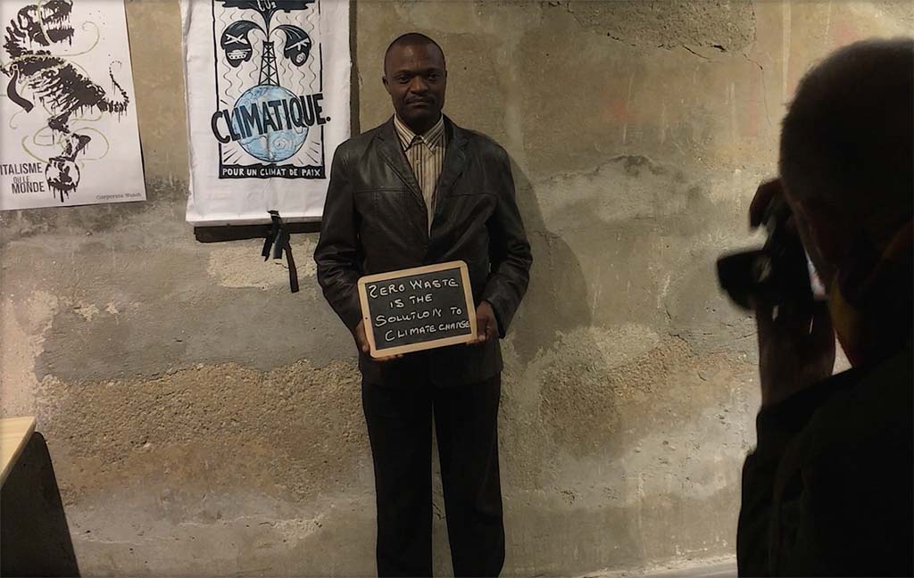 Ein Mann steht vor einer Wand und hält eine Kreidetafel mit der Aufschrift "Zero Waste is the solution to climate change" in den Händen.