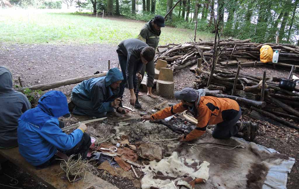 Menschen basteln aus Tierfell etwas an einer Feuerstelle im Wald.