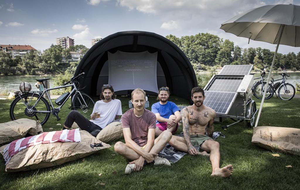 Vier Männer sitzen vor einem Zelt. Im Hintergrund steht eine mobile Solaranlage.