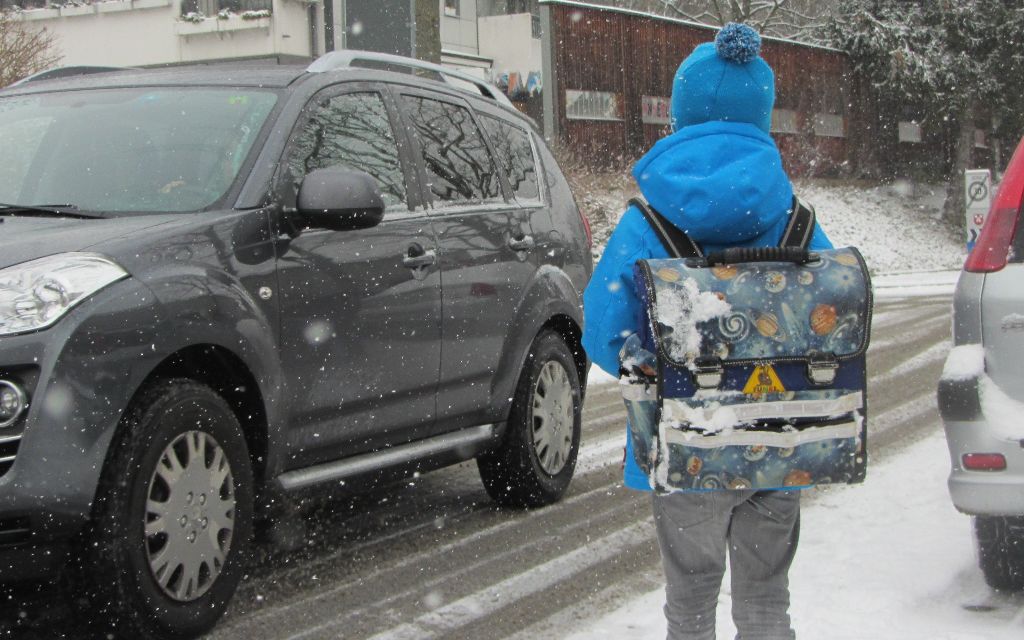 Es ist Winter und schneit. Ein Kin mit einem Schulranzen auf dem Rücken überquert eine befahrene Strasse.