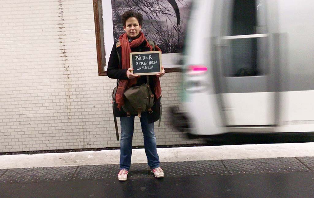 Esther Petsche steht vor einer fahrenden U-Bahn und hält eine Kreidetafel mit der Aufschrift "Bilder sprechen lassen" in den Händen.