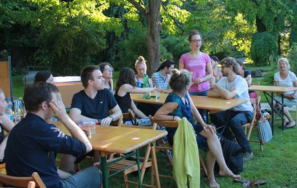 Menschen sitzen an Tischen in einem Park. Eine Frau ist am sprechen.