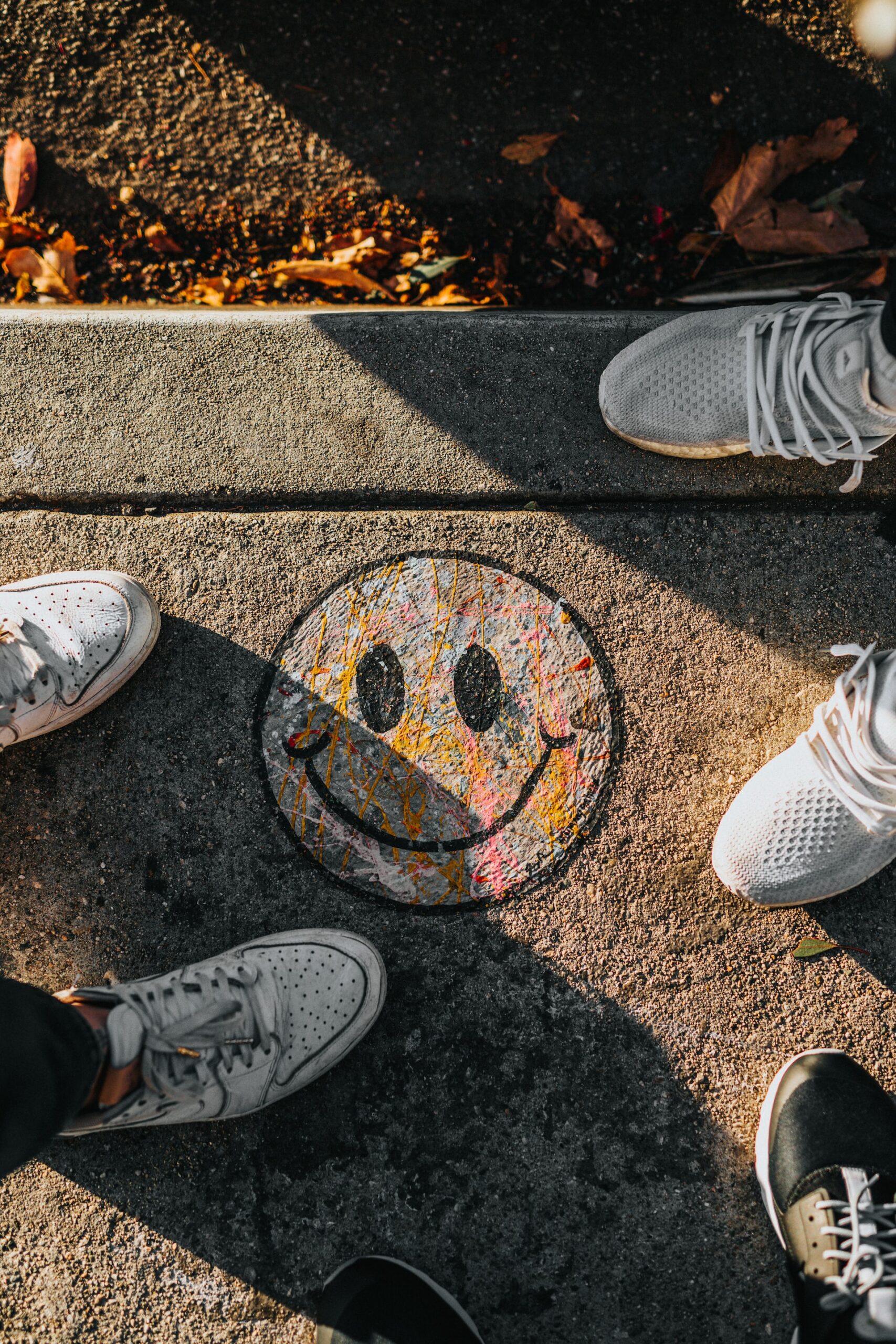 Smiley mit Kreide auf eine Strasse gemalt, drum herum Schuhspitzen.