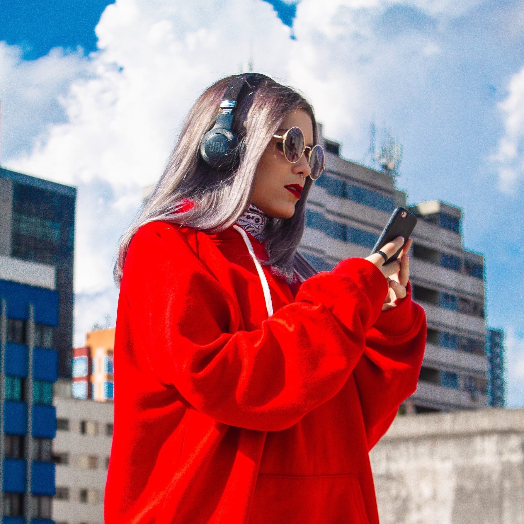 Junge Frau in rotem Pulli und zerrissener Jeans mit Kopfhörern auf dem Kopf schaut auf ihr Handy. Im Hintergrund Hochhäuser.
