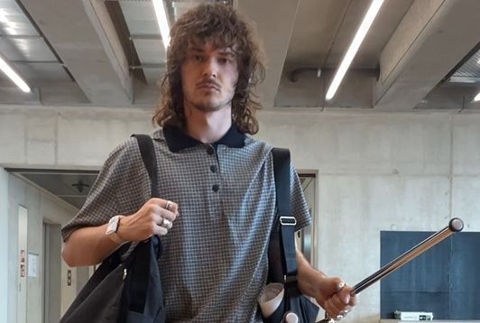 Gian hat eine Umhängetasche über die rechte Schulter und hält in der linken Hand einen Stuhl.