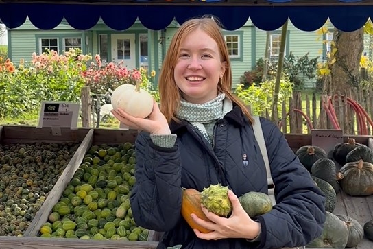 Caroline hält mehrere Kürbisse in der Hand und lächelt in die Kamera.