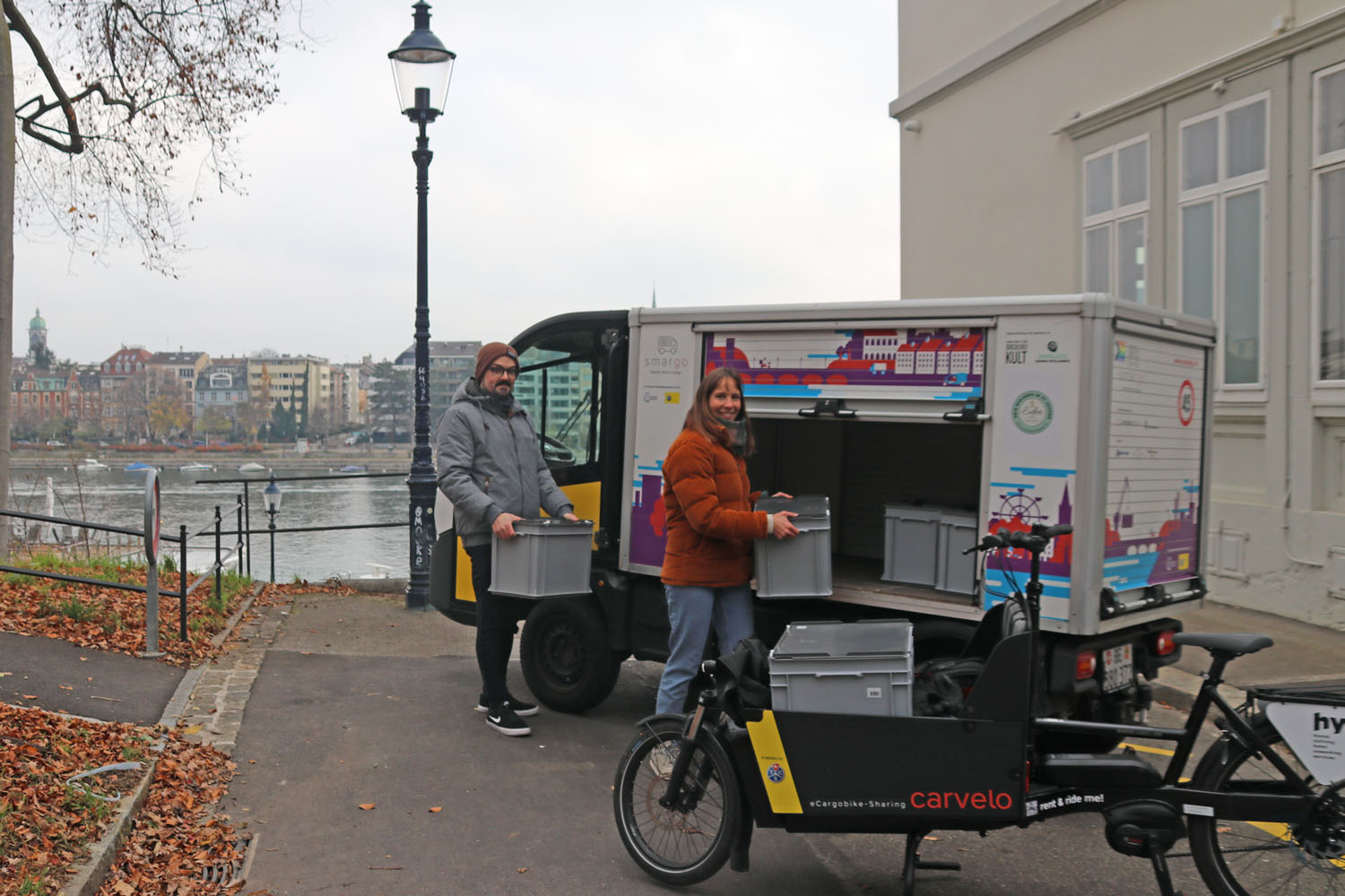 Ein Mann und eine Frau laden schwere Kisten in ein E-Transporter und ein Cargobike