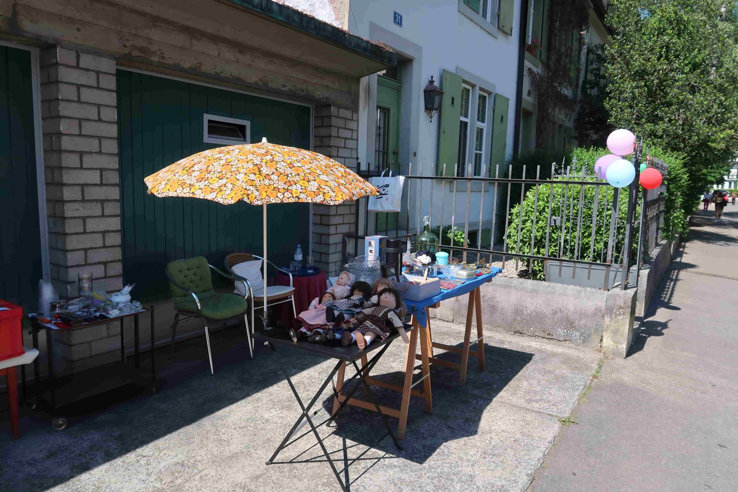 Tisch mit Flohmarktartikeln und Sonnenschirm auf Vorplatz vor Haus
