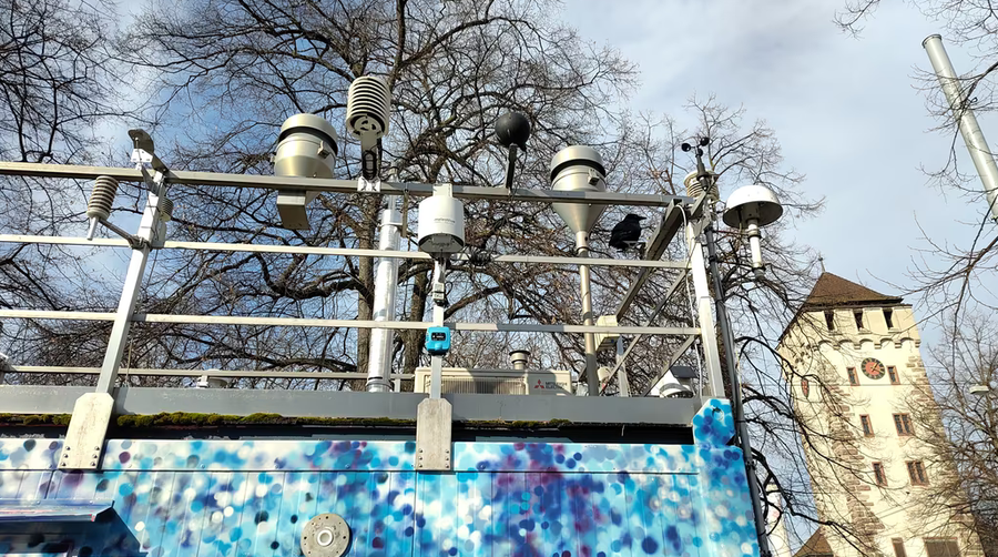 Mehrere meteorologische Geräte auf einem bunten Gebäude vor einem Turm. Messstation St. Johanns-Platz Basel.